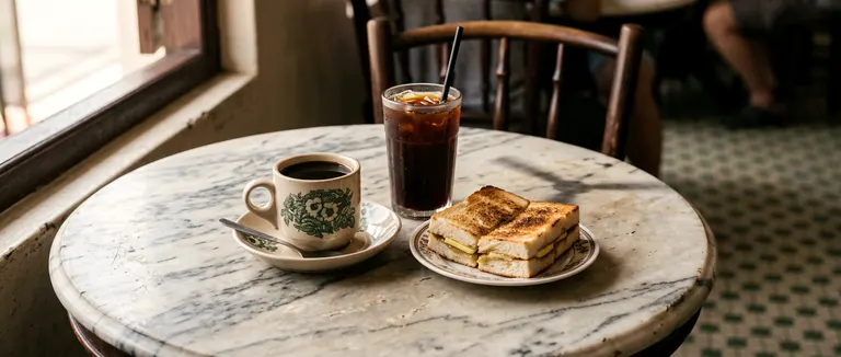 A kopitiam marble table with black coffee in a green-flowered cup, iced coffee, and kaya toast.